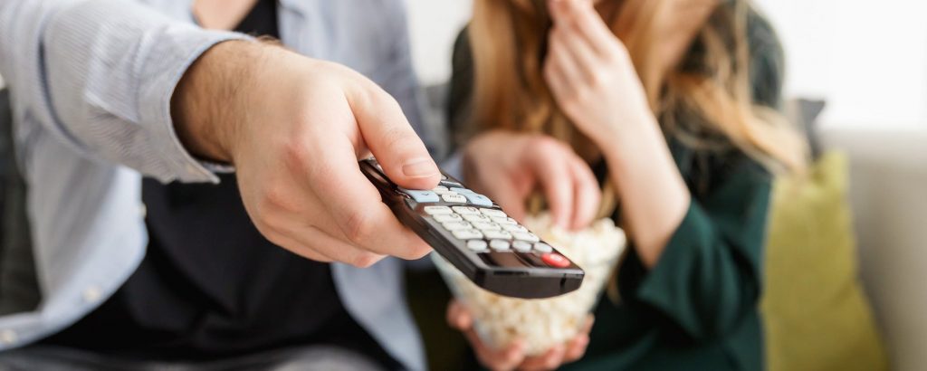 Two people sit on a sofa. One holds a TV remote in their outstretched hand. The other has a bowl of popcorn on their lap.